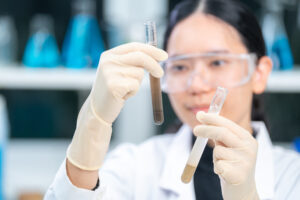 A scientist, equipped with safety goggles and gloves, conducts a lab analysis by comparing two test tubes—one containing a brown liquid and the other a pale one—set against a backdrop of blurred blue bottles in the laboratory.